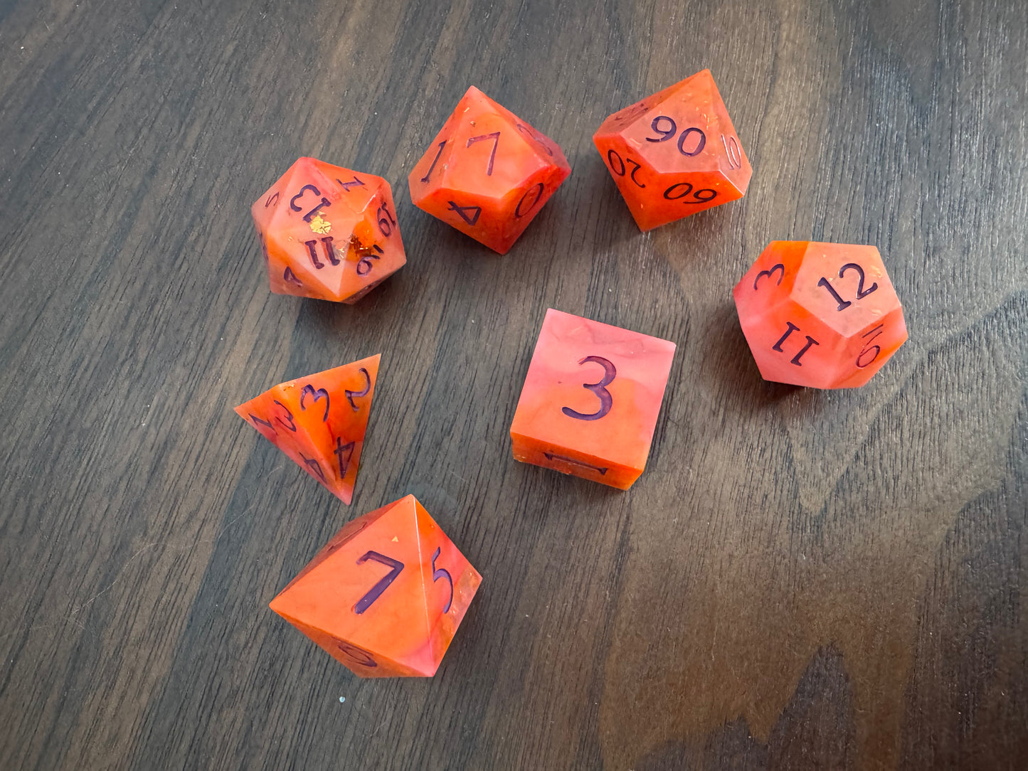 A set of 7 orange and red, swirled polyhedral dice on a wood table.