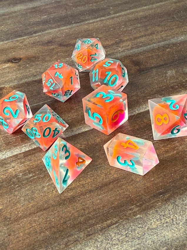 Handmade translucent polyhedral dice set with red, orange, and teal numbers on wood table