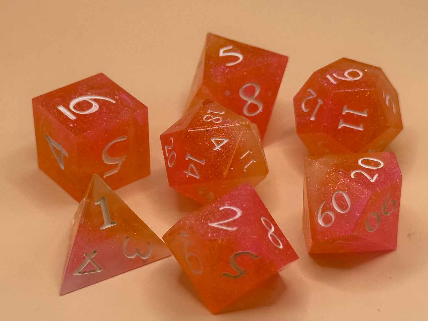 A set of seven, D&D hand-poured, resin dice in orange, pink and white on a wooden table. Numbers inked in white.