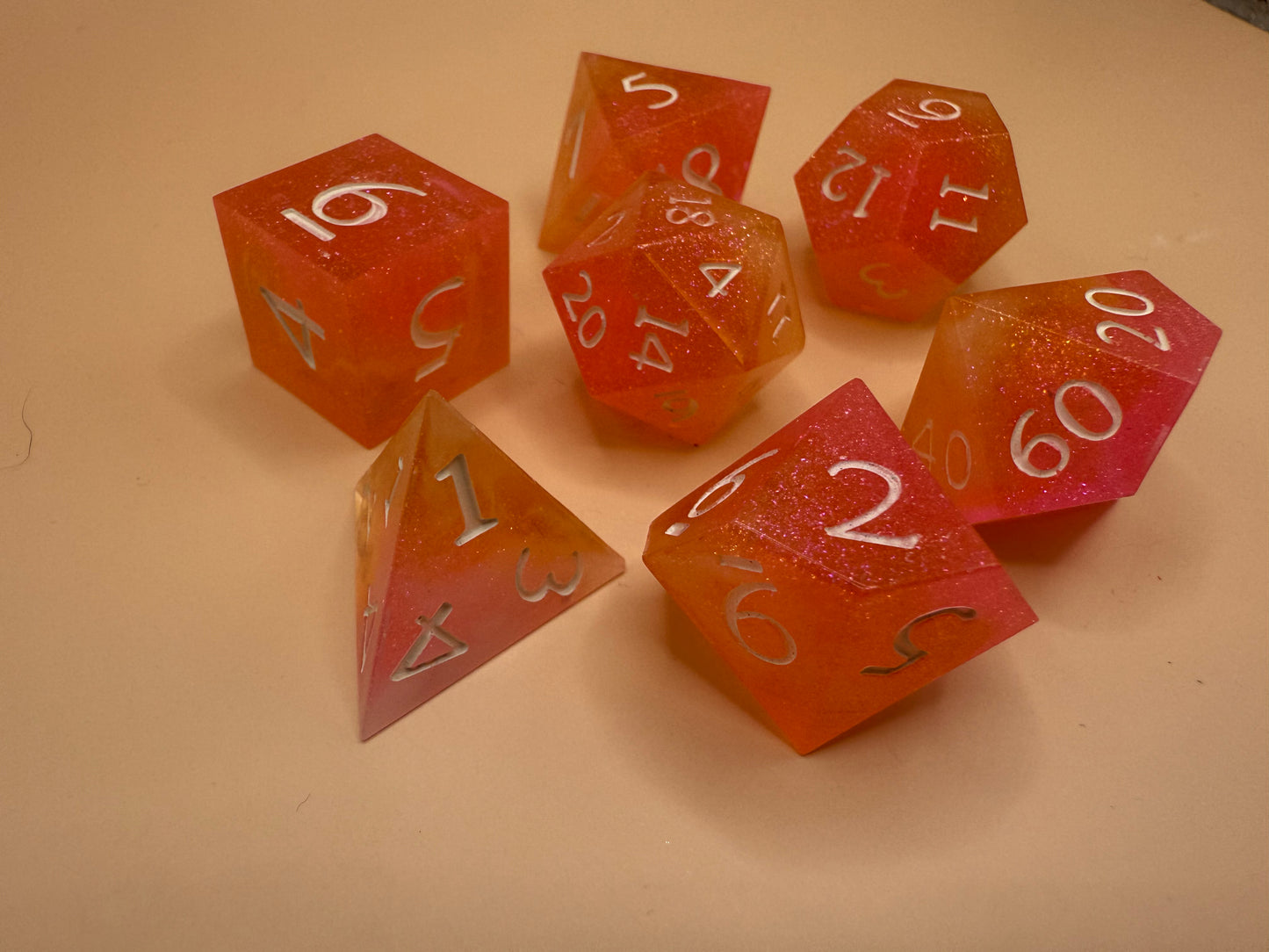 A set of seven, D&D hand-poured, resin dice in orange, pink and white on a wooden table. Numbers inked in white.