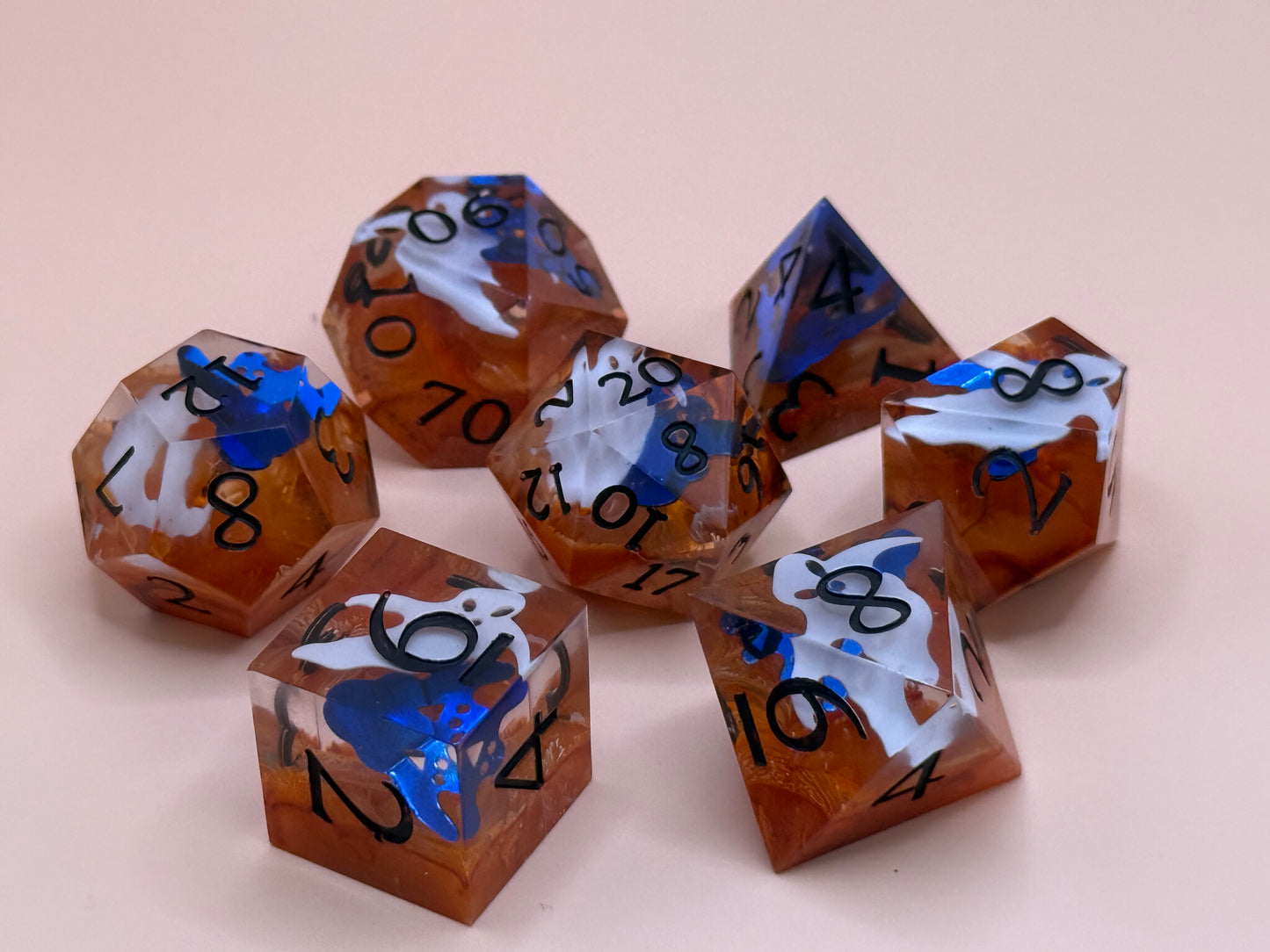 Orange sharp-sided, seven polyhedral dice with ghosts inside of them on a white surface.