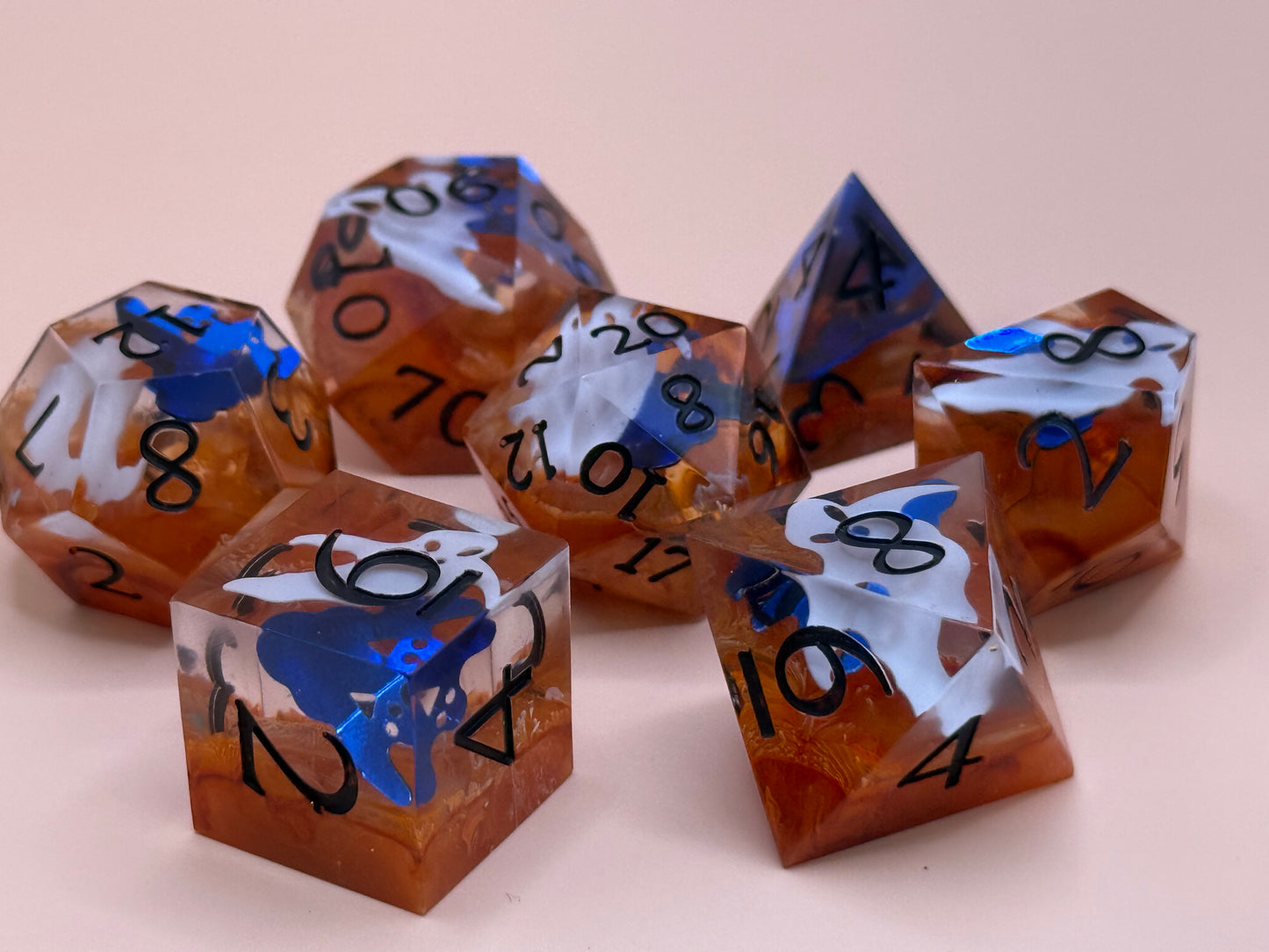 Orange sharp-sided, seven polyhedral dice with ghosts inside of them on a white surface.