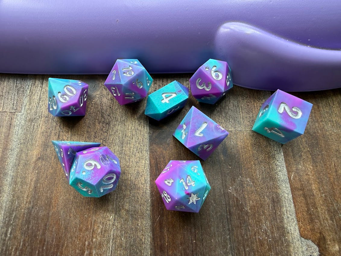 Set of purple and teal polyhedral dice on a wooden surface with a purple handle in the background.