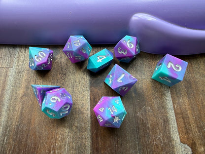 Set of purple and teal polyhedral dice on a wooden surface with a purple handle in the background.