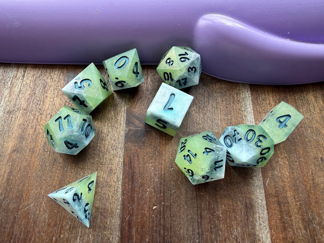 Set of green polyhedral dice on a wooden surface with a purple object in the background.