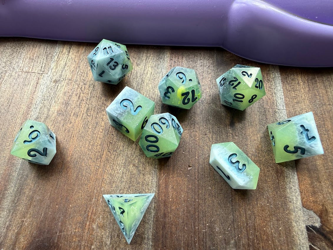 Set of green polyhedral dice on a wooden surface with a purple die in the background.