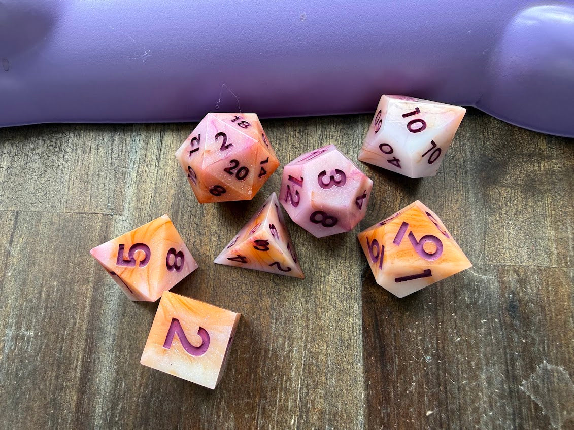 Set of colorful polyhedral dice on a wooden surface with a purple object in the background.