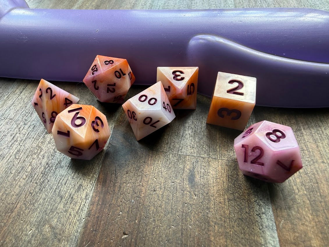 Set of polyhedral dice on a wooden surface with a purple cylindrical object in the background.