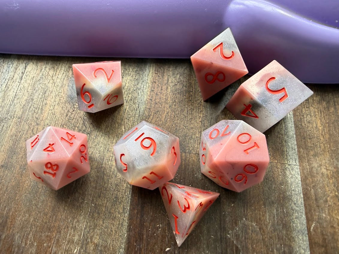 Set of pink polyhedral dice with red numbers on a wooden surface.