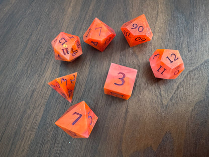 A set of 7 orange and red, swirled polyhedral dice on a wood table. 