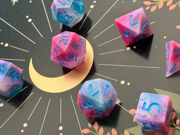 Pink and blue handmade polyhedral dice set with blue numbers on a celestial-themed background