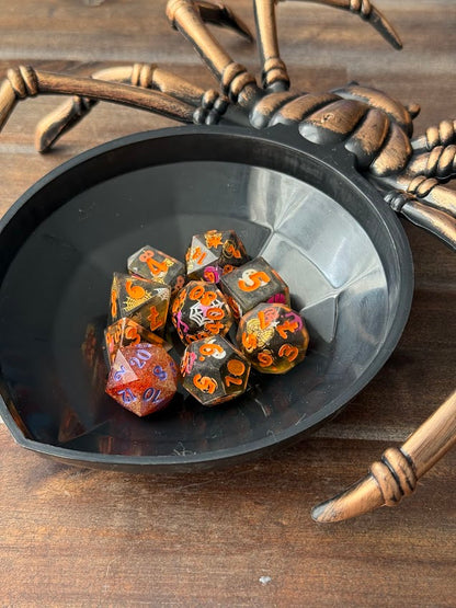 Set of dice with colorful patterns in a black dish, surrounded by a decorative spider on a wooden surface.