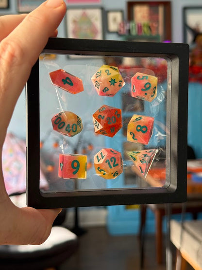 Colorful dice set in a clear plastic case held by a hand with a blurred indoor background
