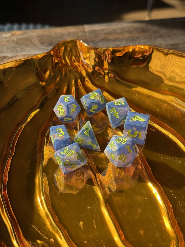 Blue resin polyhedral dice set with yellow numbers on a metallic gold tray, sunlight shining