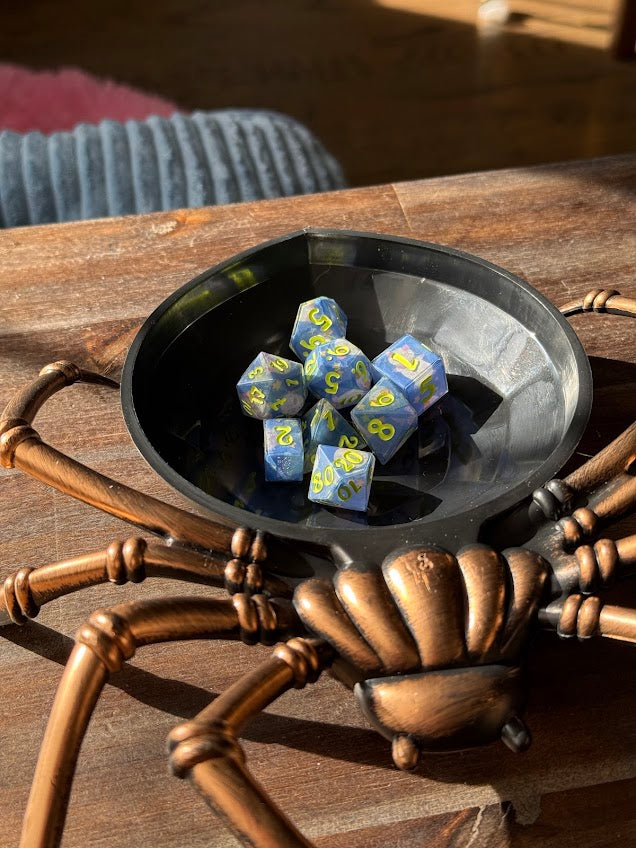 Handmade blue and yellow polyhedral dice set in a black spider-shaped dice tray on wood table