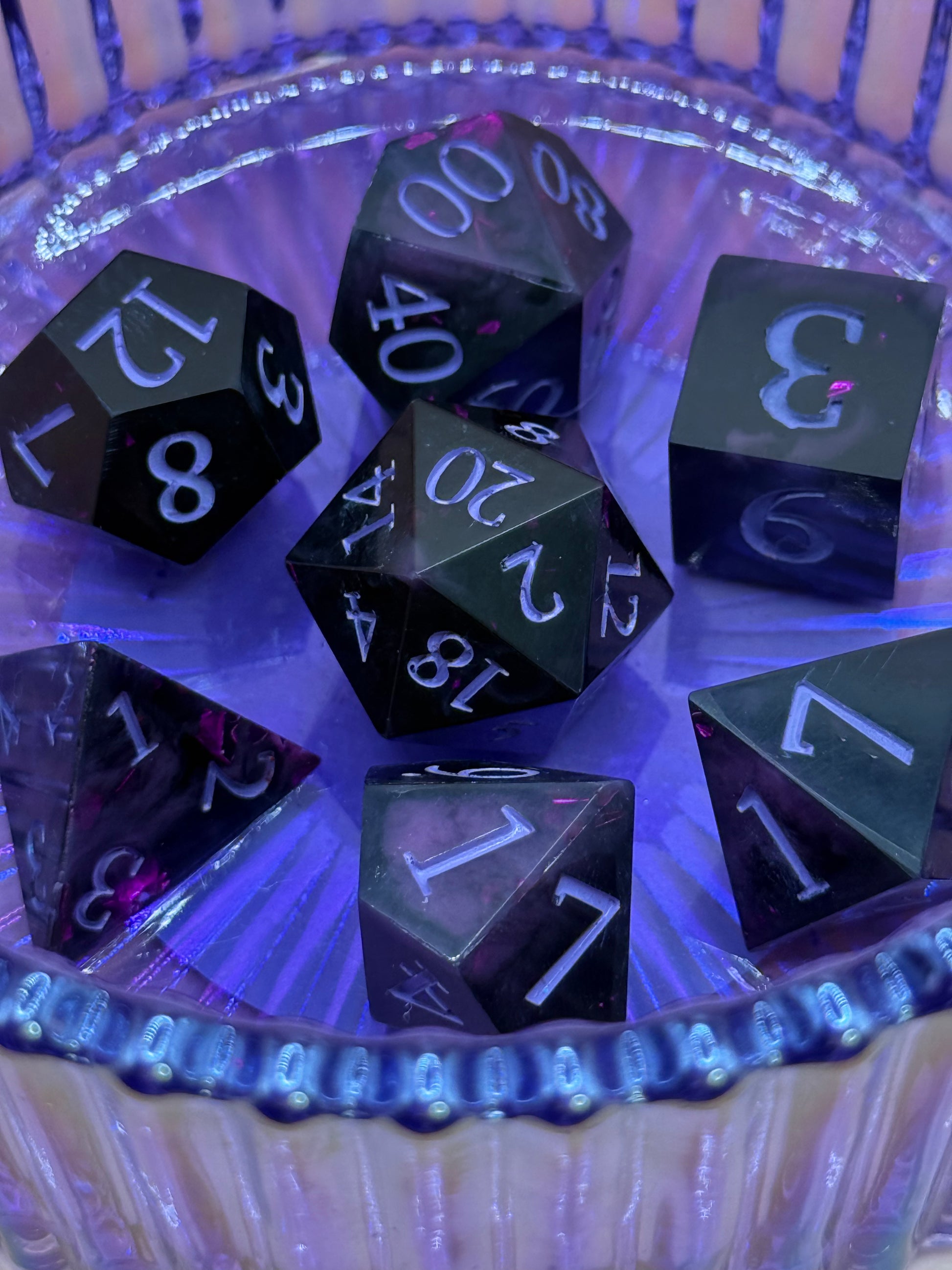 Set of black polyhedral dice with white numbers in a clear container under purple light