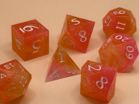 A set of seven, D&D hand-poured, resin dice in orange, pink and white on a wooden table. Numbers inked in white.