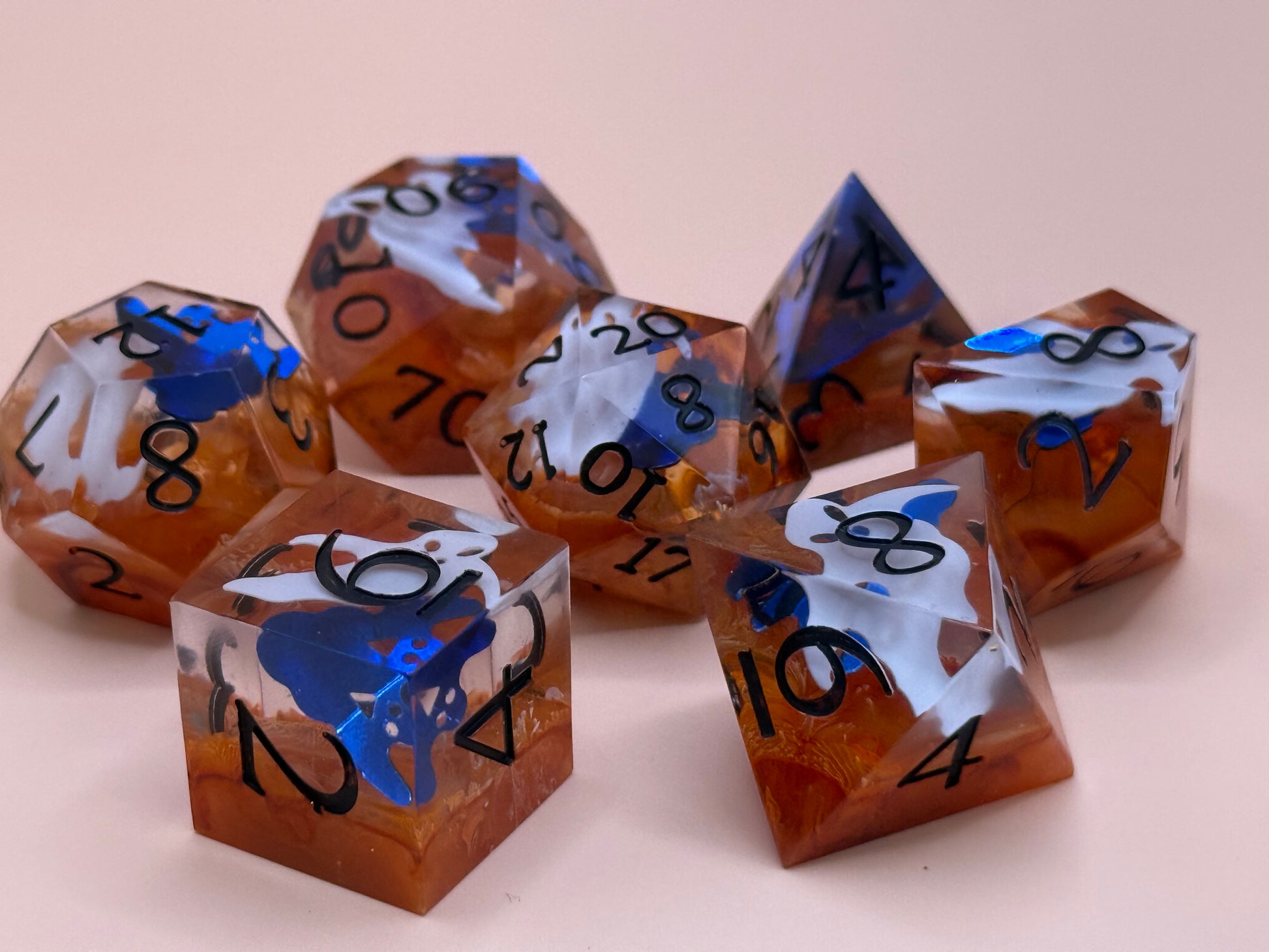 Orange sharp-sided, seven polyhedral dice with ghosts inside of them on a white surface.