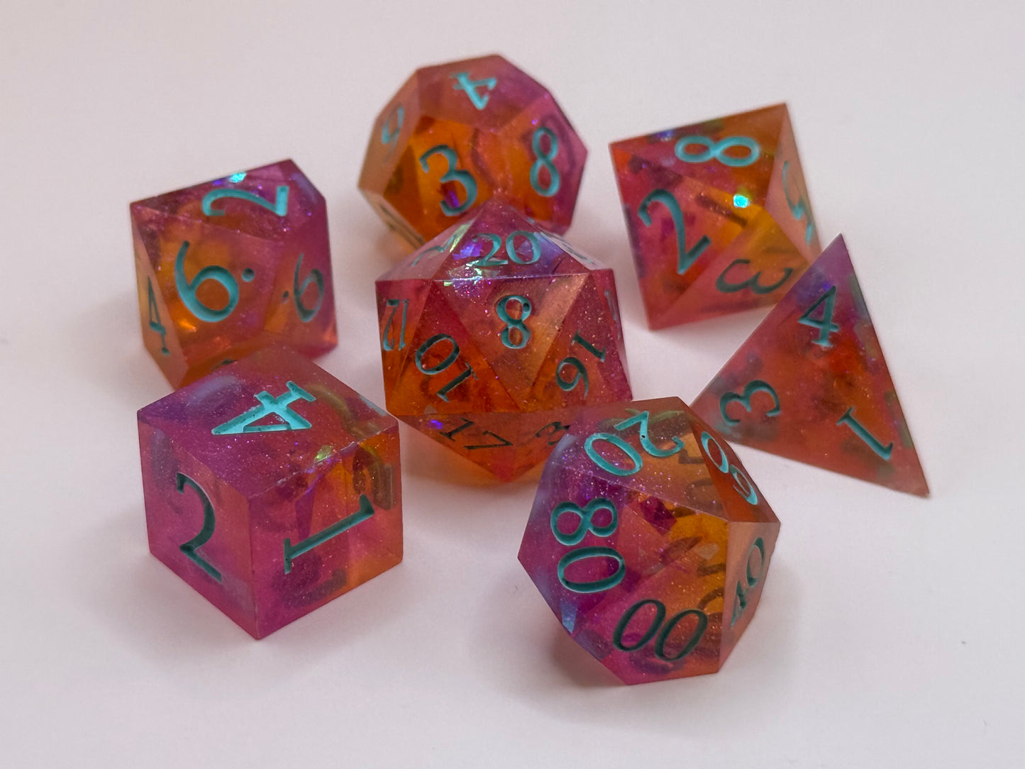 A set seven, D&D hand-poured, resin dice in orange, pink and white , with flecks of glitter, on a wooden table. Numbers inked in teal.