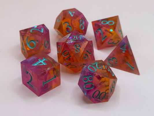 A set seven, D&D hand-poured, resin dice in orange, pink and white , with flecks of glitter, on a wooden table. Numbers inked in teal.