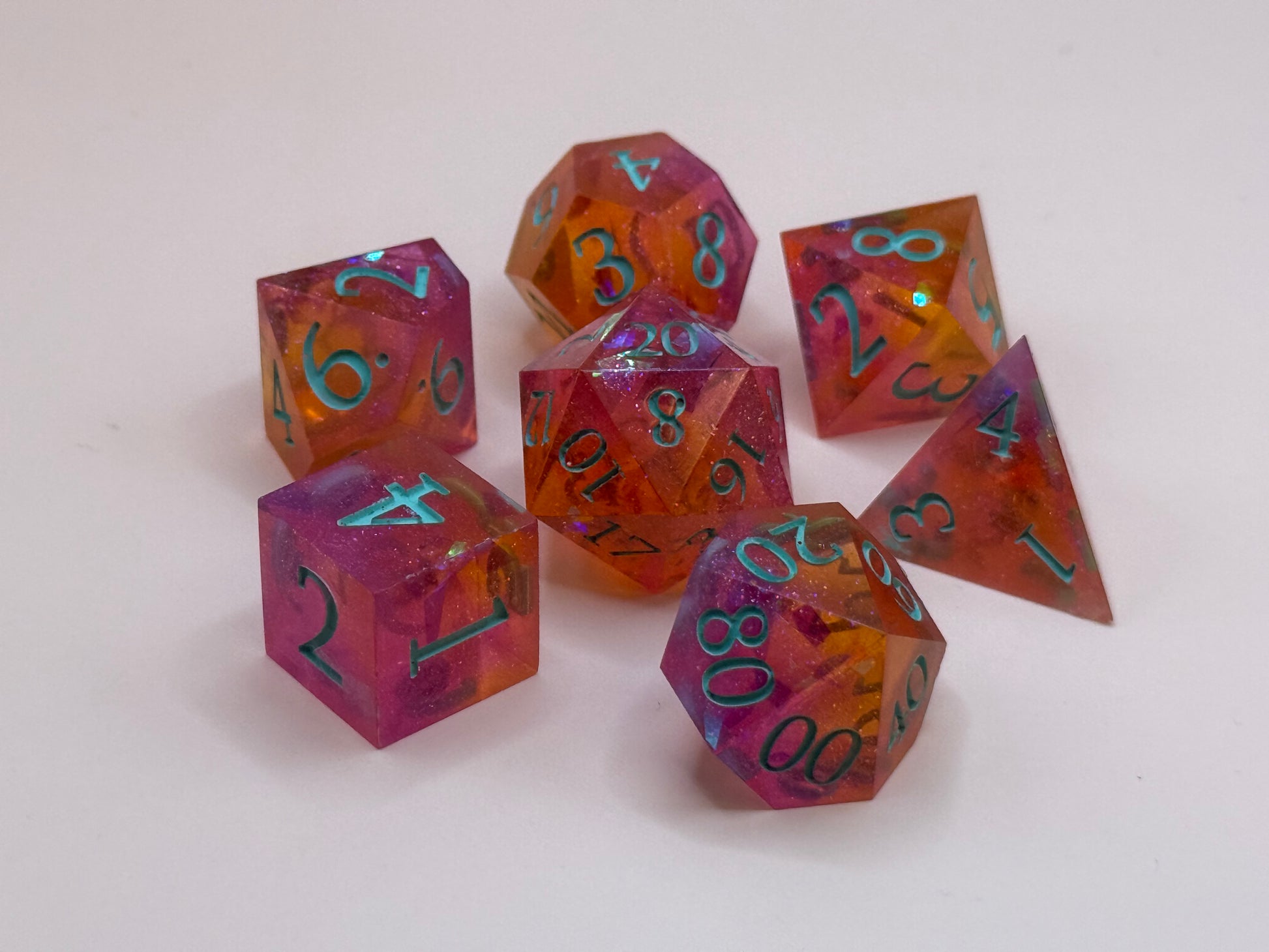 A set seven, D&D hand-poured, resin dice in orange, pink and white , with flecks of glitter, on a wooden table. Numbers inked in teal.