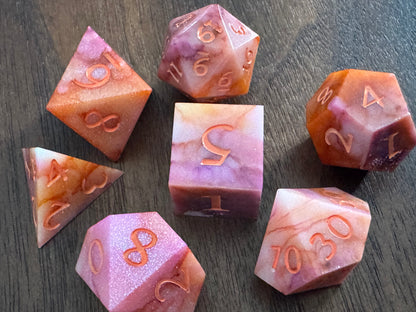 Orange, pink, and white, marbled sharp-edge hand-poured resin dice displayed on a table. 