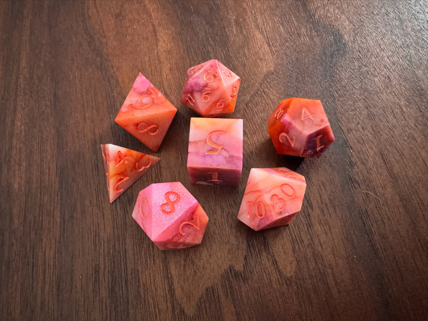 Orange, pink, and white, marbled sharp-edge hand-poured resin dice displayed on a table. 