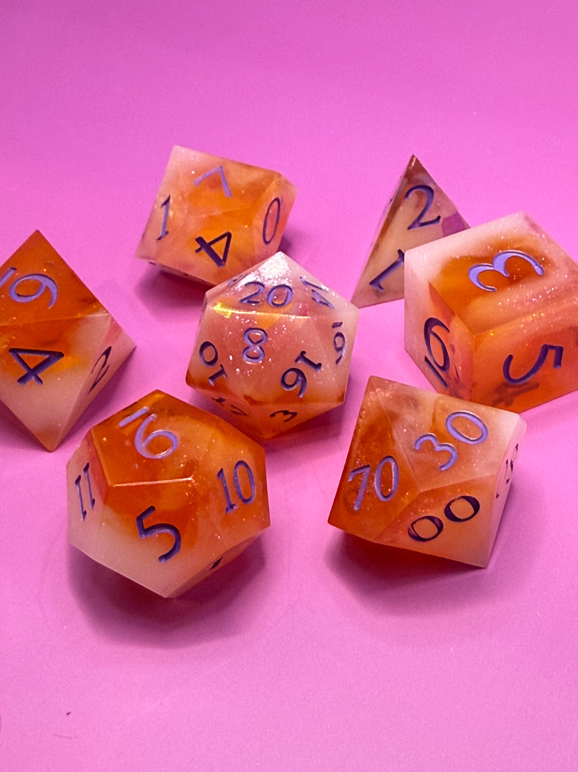 Seven hand-poured orange, pink, and white D&D resin dice on a wood table. Inked in purple. 