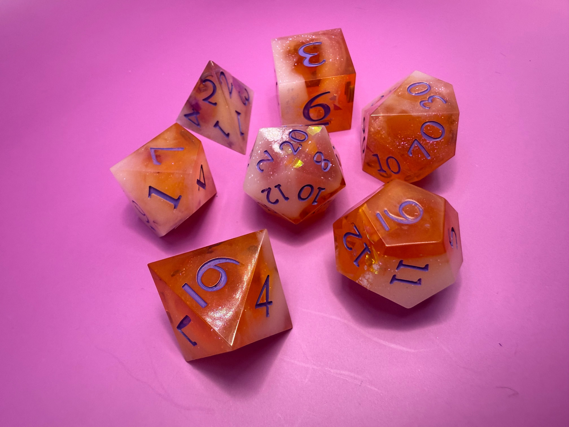 Seven hand-poured orange, pink, and white D&D resin dice on a wood table. Inked in purple. 
